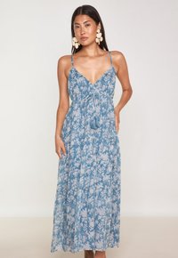 Robe maxi bleu clair avec un motif floral blanc. Elle possède un décolleté en V profond, des bretelles ajustables et un détail de lien avec des pompons à la taille.
