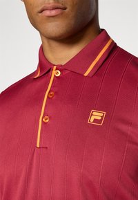 Maroon polo shirt with horizontal stripes, two orange accent stripes on the collar, four orange buttons, and a small yellow logo on the chest.