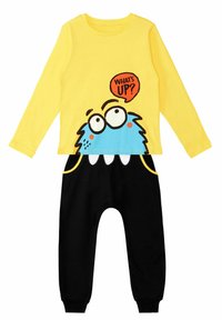 Gelbes Langarmhemd mit einem blauen Monster-Motiv und dem Text "Was ist los?", kombiniert mit schwarzen Hosen mit gelben Akzenten und Taschen.