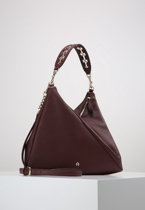 Donkerbruine leren handtas met een driehoekige vorm, kettinghandvat, ritssluitingvak en verstelbare schouderband, tentoongesteld op een wit platform.