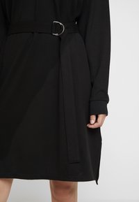 Robe noire en tissu doux, dotée d'une taille ceinturée avec une boucle circulaire en argent, des manches longues et un ourlet droit avec des fentes latérales.