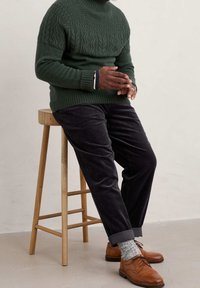 Dunkelgrüner Kabelstrickpullover mit hohem Kragen, kombiniert mit dunkelbraunen Cordhosen. Braune Lederschuhe und gemusterte Socken sind sichtbar.