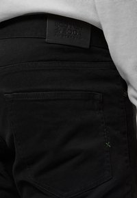 Μαύρο τζην παντελόνι με δερμάτινη ετικέτα «Scotch & Soda» στη μέση και τσέπη με πράσινη ραφή.