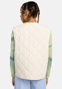 Gilet matelassé crème avec un motif en diamant. Présente des bords arrondis et est superposé sur une chemise à manches longues colorée à motifs.