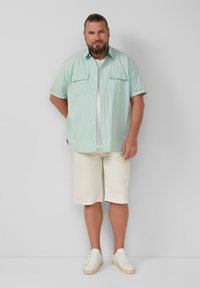 Himmelblaues kurzärmliges Hemd mit Knopfleiste und zwei Brusttaschen, kombiniert mit cremefarbenen knielangen Shorts und weißen Turnschuhen.