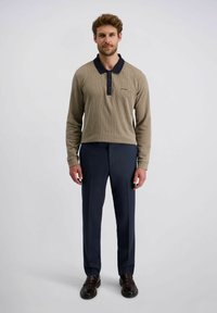 Man staat in een beige lange mouwen polo met zwarte kraag, marineblauwe broek en donkerbruine nette schoenen tegen een effen achtergrond.