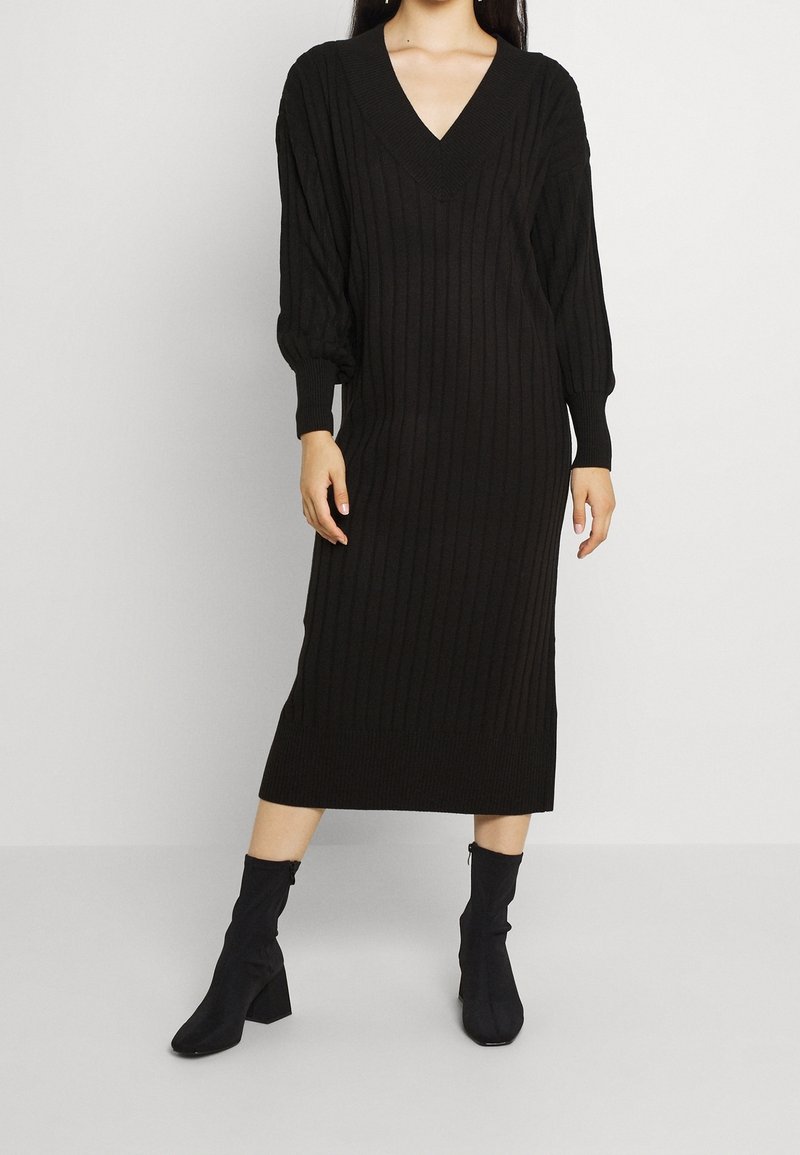 Abito nero lavorato a maglia con texture a coste, scollatura a V profonda e maniche lunghe a sbuffo. Abbinato a stivali neri alla caviglia con tacco spesso.