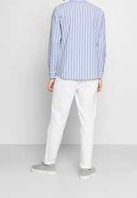 Osoba ubrana w jasnoniebieską i białą pionową w paski koszulę z długim rękawem, białe spodnie i szare buty slip-on, stojąca tyłem do aparatu.