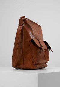 Brun læder skuldertaske med en frontlomme og justerbar rem, præsenteret på en hvid piedestal mod en grå baggrund.