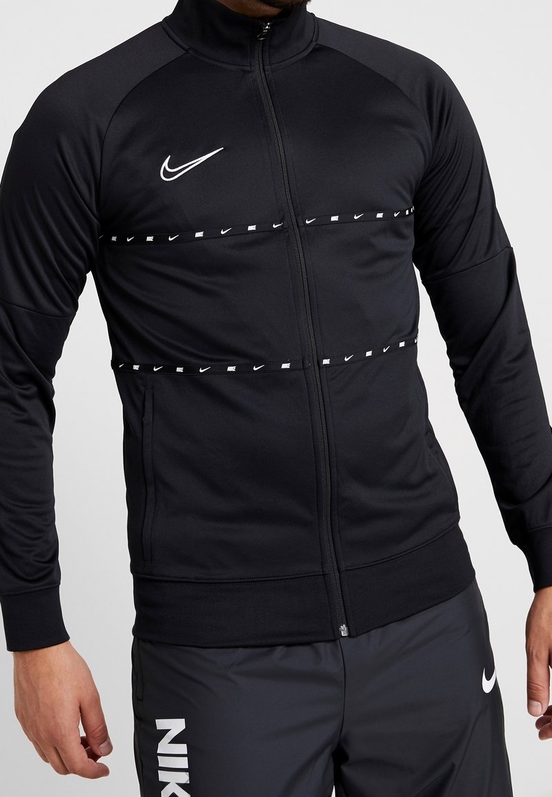 Veste noire Nike zippée avec logo blanc et bande horizontale de marque sur la poitrine, portée avec un pantalon Nike foncé montrant des logos partiels.