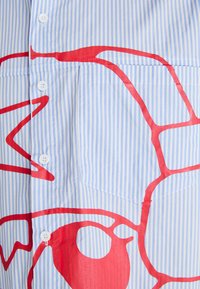 Chemise boutonnée rayée bleu clair et blanche avec un grand motif abstrait rouge ; elle comporte une poche poitrine et des boutons blancs.