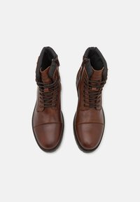 Bottes chevilles en cuir marron avec des accents cousus, bout rond, à lacets à l'avant et fermeture éclair. Doublure en textile texturé, semelle en caoutchouc.