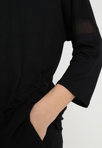 Schwarzes Oberteil mit Dreiviertelärmeln, aus weichem Material. Transparente Details an den Ärmeln, lässiger Schnitt, seitliche Tasche sichtbar.