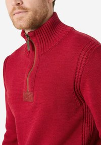 Roter gestrickter Pullover mit hohem geripptem Kragen, kurzem Reißverschluss und strukturierten Seitenpanelen. Mit einem Wildlederakzent am Reißverschluss.