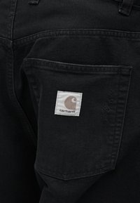 Τα μαύρα τζιν παντελόνια διαθέτουν πίσω τσέπη με ένα τετράγωνο λευκό ετικέτα που εμφανίζει ένα λογότυπο. Το ύφασμα έχει λεία υφή με ορατή ραφή.