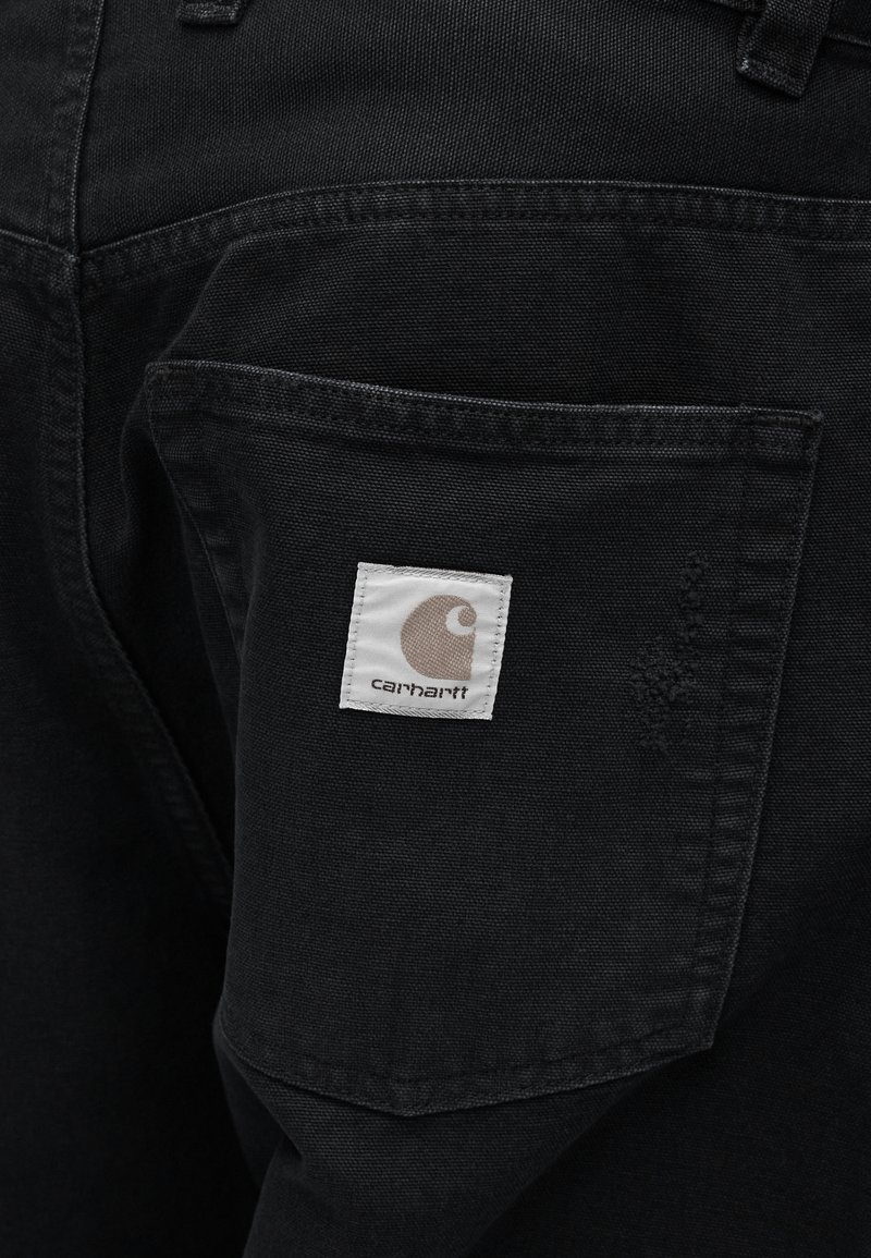 Sorte denimbukser har en baglomme med et kvadratisk hvidt mærke, der viser et logo. Stoffet har en glat tekstur med synlige syninger.