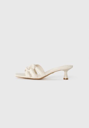 Crème-kleurige schuifsandalen met drie gestructureerde banden en een geknoopt detail. Ze hebben een lage, taps toelopende hak met een lichte bruine accent.