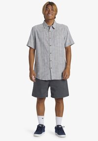 Kurzärmliges Hemd in Hellgrau mit kariertem Muster, Tasche, kombiniert mit grauen Shorts und marineblauen Sneakers, weiße Socken sichtbar.