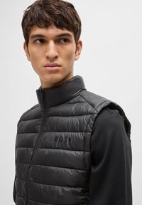 Black padded vest with high collar, quilted texture, front zipper, and “BOSS” logo on the chest. Worn over a black long-sleeve shirt.