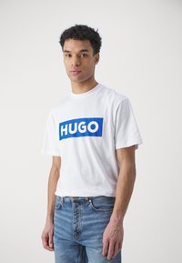 Tricou alb din bumbac, cu un grafic rectangular albastru pe care este scris "HUGO" cu litere albe îngroșate. Asortat cu blugi albaștri.