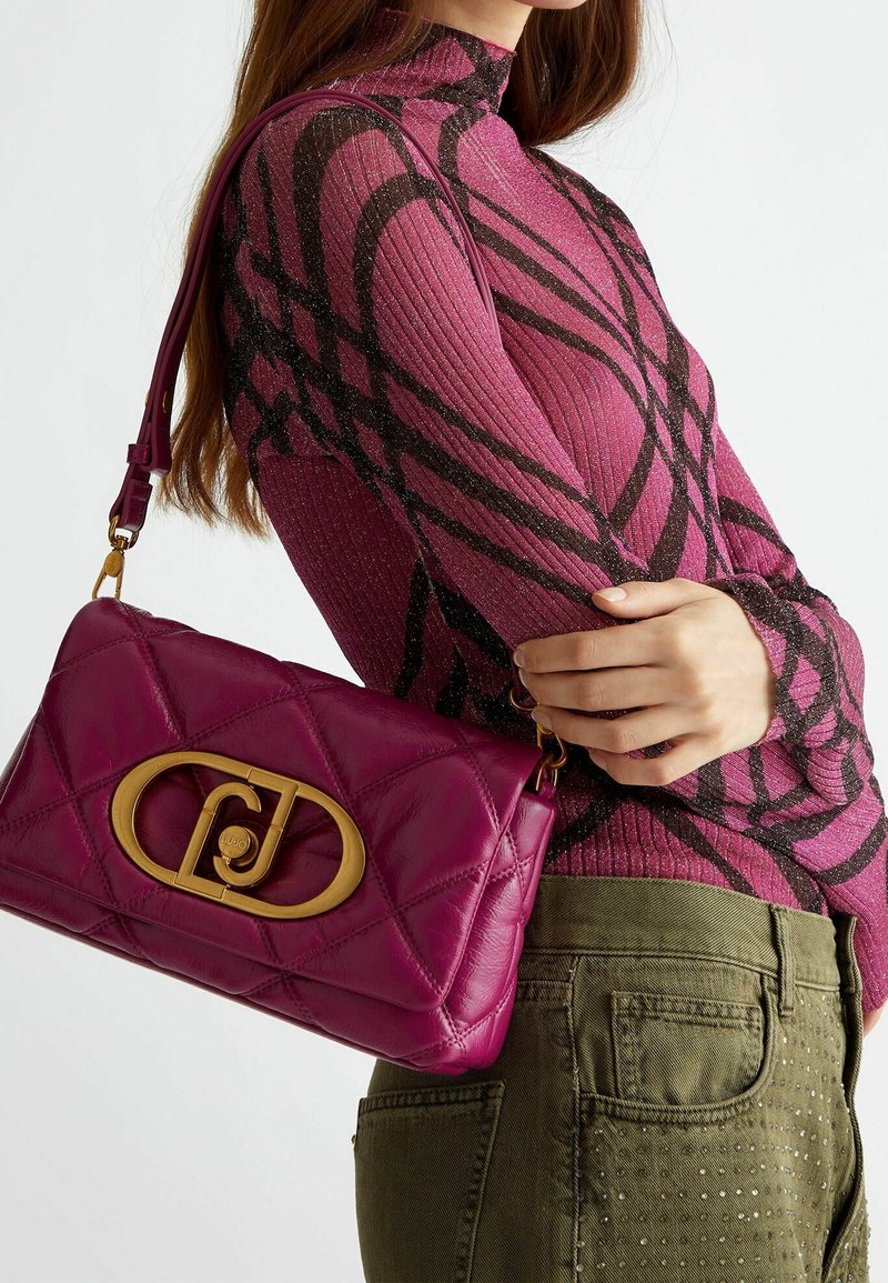 Femme portant un pull en maille côtelée rose à motifs et un pantalon olive, portant un sac à bandoulière matelassé magenta avec une grande boucle dorée.