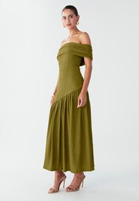 Olivengrøn skulderfri kjole med tekstureret finish, tætsiddende overdel og detaljeret, rynket nederdel; kombineret med nøgne hælede sandaler.
