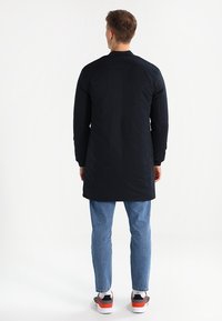 Cappotto lungo nero con una texture liscia, caratterizzato da un taglio dritto e un colletto alto, abbinato a jeans azzurri e scarpe sportive.