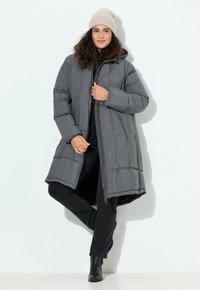Graue Pufferjacke mit lockerer Passform, ausgestattet mit einem Reißverschluss vorne, einem gesteppten Muster und einem mittellangen Design. Getragen über schwarzer Kleidung und Stiefeln.