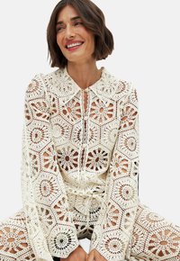 Top in cotone bianco con intricate lavorazioni a rombo e floreali, colletto e chiusura con bottoni. Maniche lunghe e dettagli a punto aperto.