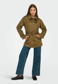 Brun mocka jacka med krage, framknappning och bälte, matchad med vida blå denimjeans med pärldekorationer.