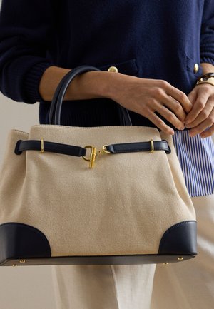 Μπεζ τσάντα tote με μαύρες λαβές, μαύρες δερμάτινες λεπτομέρειες και χρυσά μεταλλικά στοιχεία, που κρατιέται από ένα άτομο που φοράει μπλε σκούρο πουλόβερ και ριγέ πουκάμισο.