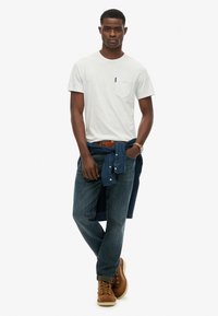 Witte T-shirt met korte mouwen en een borstzak, blauwe jeans en bruine laarzen. Een blauw hemd is om de taille geknoopt. Eenvoudige, casual outfit.