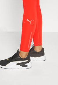 Nogi w jasnoczerwonych legginsach Puma i czarnych sneakersach Puma z białymi podeszwami na jednolitym tle.