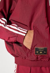 Casaco corta-vento grená com riscas brancas nas mangas, punhos elásticos e barra elástica. Apresenta uma etiqueta de tecido com detalhes do logótipo no bolso frontal.