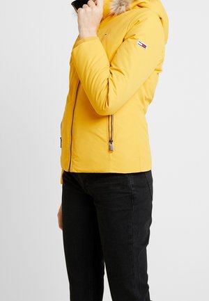 Veste rembourrée jaune avec une capuche en fourrure, poches zippées et un accent de logo sur la manche. Texture lisse et design ajusté.