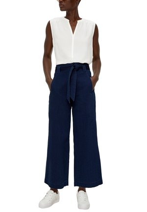 Donna che indossa una camicetta bianca senza maniche, pantaloni navy a gamba larga a vita alta con cintura annodata, mani nelle tasche e sneakers bianche.