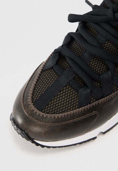 Zapatilla deportiva de malla negra y cuero marrón con puntera redonda, cordones negros gruesos y suela de goma texturizada, con detalles de costura en contraste.