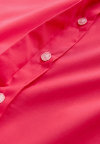 Jasnoróżowa tkanina o gładkiej fakturze, z trzema plastikowymi guzikami z przodu. Prosty, klasyczny fason z zapięciem na guziki.