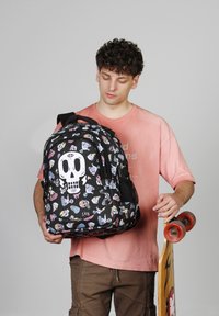 Mochila negra con gráficos coloridos de calaveras y temática de skate, con correas ajustables y bolsillos de malla laterales. Hombre sosteniendo una patineta.