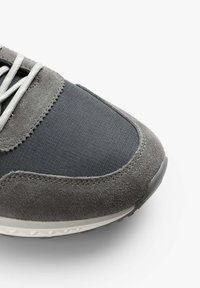 Sneaker grigio realizzato con una combinazione di materiali in suede e tessuto, dotato di lacci rotondi, superficie testurizzata e suola in gomma.