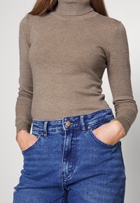 Lichtbruine coltrui met een geribbelde textuur, gecombineerd met hoog-getailleerde, vervaagde blauwe denim jeans. Zakjes zichtbaar op de taille.