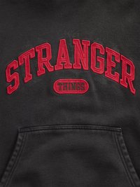 Μαύρο φούτερ με κόκκινα κεντημένα γράμματα «STRANGER THINGS» που σχηματίζουν τόξο πάνω από ένα μικρό οβάλ με το ίδιο κείμενο. Περιλαμβάνει τσέπη μπροστά.
