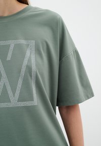 Olivza zaļš pārāk liels t-krekls ar īsām piedurknēm, ar baltu punktētu taisnstūra logotipa dizainu uz priekšpuses. Mīksta, gluda auduma.