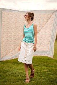 Femme portant des lunettes de soleil, un débardeur rayé turquoise et une jupe blanche, marchant sur l’herbe avec un tissu à motifs en arrière-plan en extérieur.