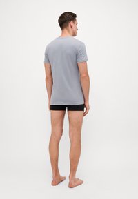T-shirt gris à manches courtes, col rond, tissu doux. Modèle vu de dos, portant un short noir, pieds nus. Design simple.