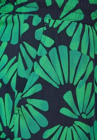 Tessuto verde e nero con un motivo floreale caratterizzato da forme stilizzate, principalmente in verde brillante su uno sfondo scuro. Dettaglio della vita elastica.