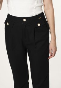 Schwarze Hose mit hohem Bund, mit goldenen Knopfakzenten und gefalteten Fronttaschen, aus einem glatten Stoff gefertigt.