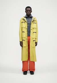 Sárga vízálló kabát több zsebbel, szürke magas nyakú felső és narancssárga széles szárú nadrág fölé viselve, fekete cipőkkel párosítva.