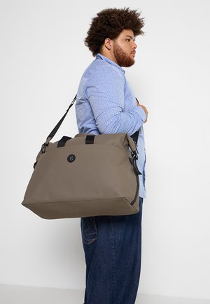 Donker taupe duffel tas met zwarte accenten, met een bovenste sluiting en een verstelbare schouderriem. Gemaakt van duurzaam materiaal met een gladde textuur.