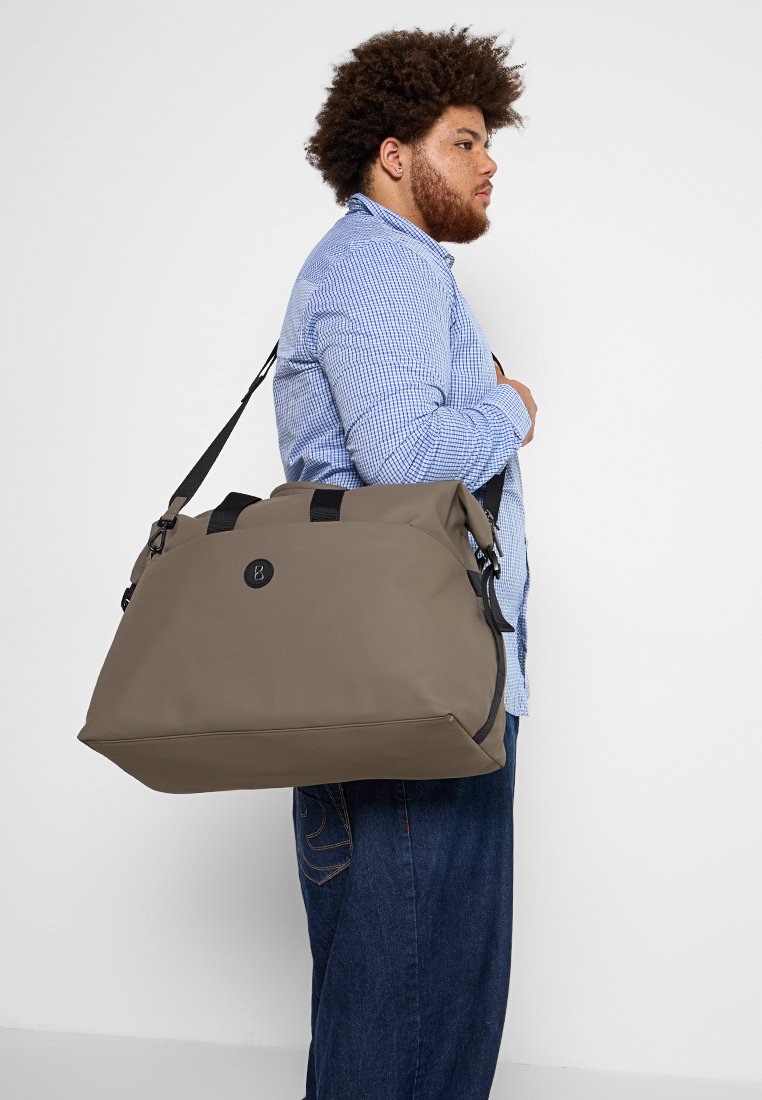 Temno taupe potovalna torba z črnimi detajli, s stranskim zapiranjem in nastavljivim ramenskim trakom. Izdelana iz trpežnega materiala z gladko teksturo.